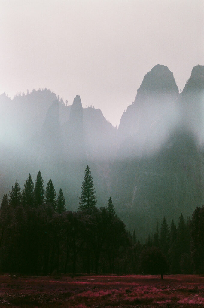 Photograph of the Cathedral Rocks in Yosemite shrouded in smoke. The treeline in the distance mimics the shapes of the rock formation, and the foreground consists of deep red foliage.