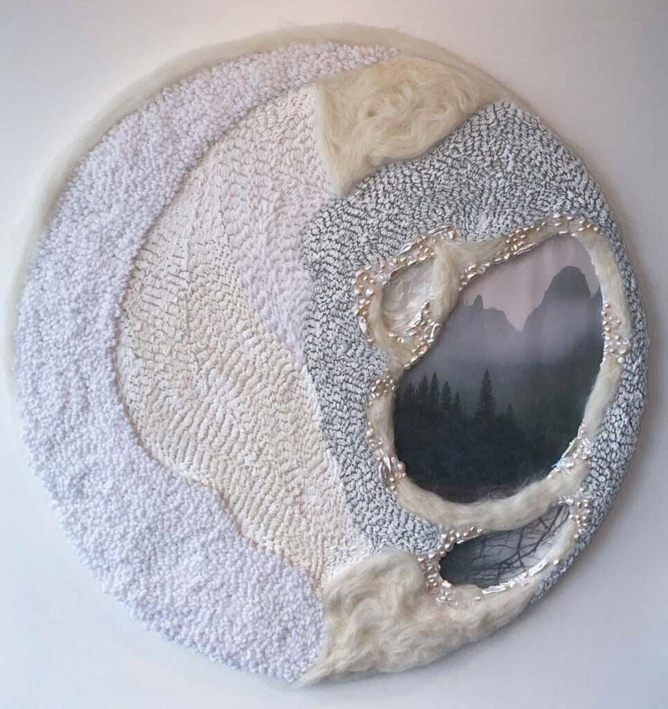 Circular punch-needle sculpture/wall hanging. A variety of yarn and wool create abstract shapes, referencing different structures in the brain. To the right of the object there are three holes bordered by pearls and translucent beads; inside the holes are different natural landscapes. The sculpture follows a monochromatic color palette of whites and grays.